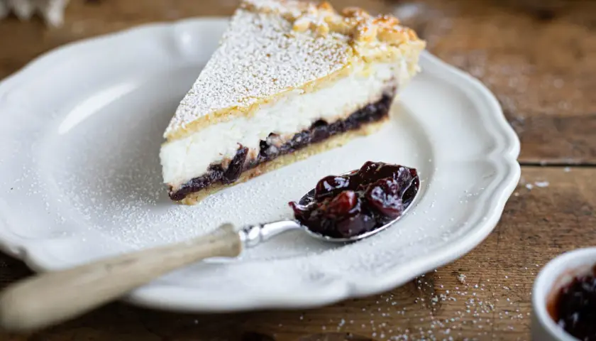 Una fetta di crostata ricotta e visciole, cosparsa di zucchero a velo, su un piatto bianco. Un cucchiaino pieno di visciole confettate accanto.