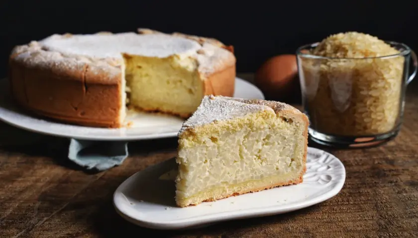 Una fetta di torta di riso, soffice e cosparsa di zucchero a velo, accanto alla torta intera e a una tazza di riso crudo.