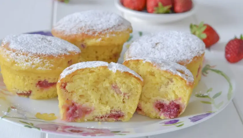 Morbidi muffin alle fragole, cosparsi di zucchero a velo, su un piatto fiorito. Uno è tagliato a metà, rivelando il ripieno succoso.