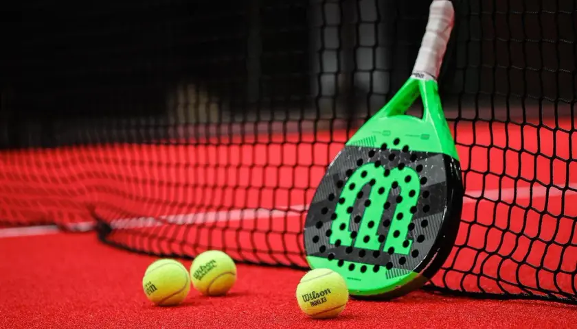 Racchetta da padel verde e nera appoggiata alla rete, con palline gialle sul campo rosso. Quanto dura una racchetta da padel? Dipende dall'uso!