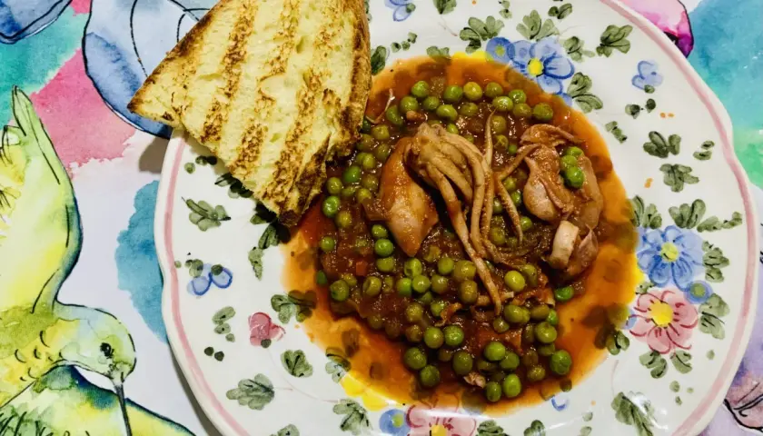 Piatto di totani con piselli in umido, accompagnato da una fetta di pane abbrustolito, su un piatto decorato con fiori e uccelli.