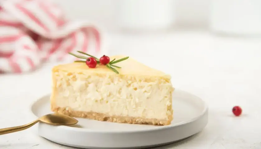 Una fetta di cheesecake con yogurt greco, guarnita con rosmarino e bacche rosse, su un piatto bianco con cucchiaino dorato.