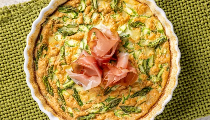 Frittata di asparagi al forno, dorata e invitante, guarnita con fette di prosciutto crudo. Un piatto perfetto per un pranzo leggero.