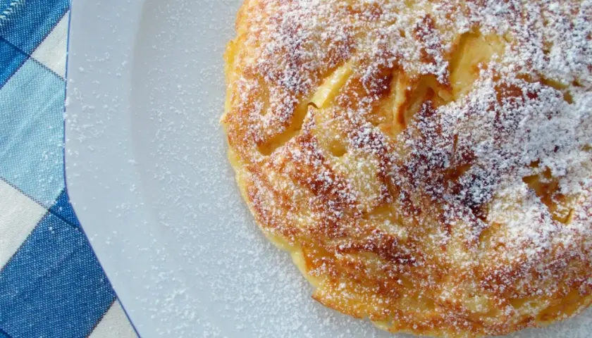 Frittata di mele in padella pronta in 5 minuti, cosparsa di zucchero a velo, su un piatto bianco accanto a una tovaglia a quadri blu.