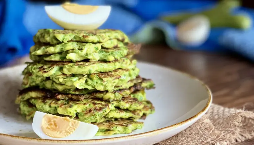 Una pila di rosti di zucchine dorati, guarniti con spicchi di uovo sodo, su un piatto bianco.