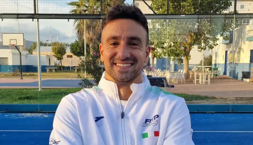 Aris Patiniotis sorride con le braccia conserte su un campo da padel blu. Indossa una giacca bianca con logo e bandiera italiana.