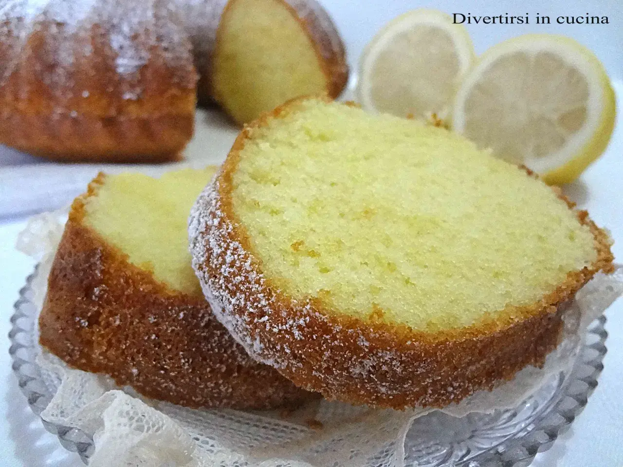 Fette di ciambellone al limone spolverate di zucchero a velo, con spicchi di limone sullo sfondo.