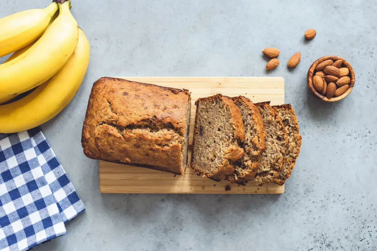 Un delizioso banana bread appena sfornato, affettato su un tagliere di legno, con banane mature e mandorle sparse.