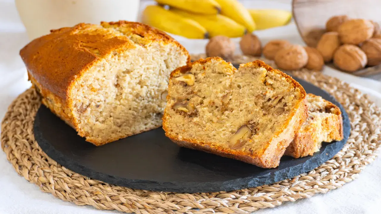 Fetta di banana bread senza burro, soffice e ricca di noci, con banane mature e noci sullo sfondo.