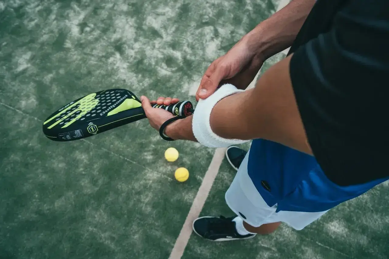 Giocatore di padel controlla la misura impugnatura racchetta padel, pronto per il servizio. Due palline gialle a terra.