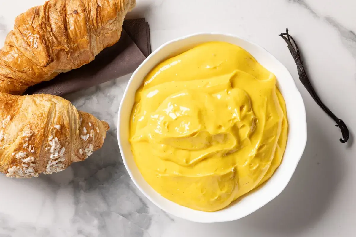 Una ciotola di cremosa crema pasticcera senza glutine, con accanto due croissant e un baccello di vaniglia.