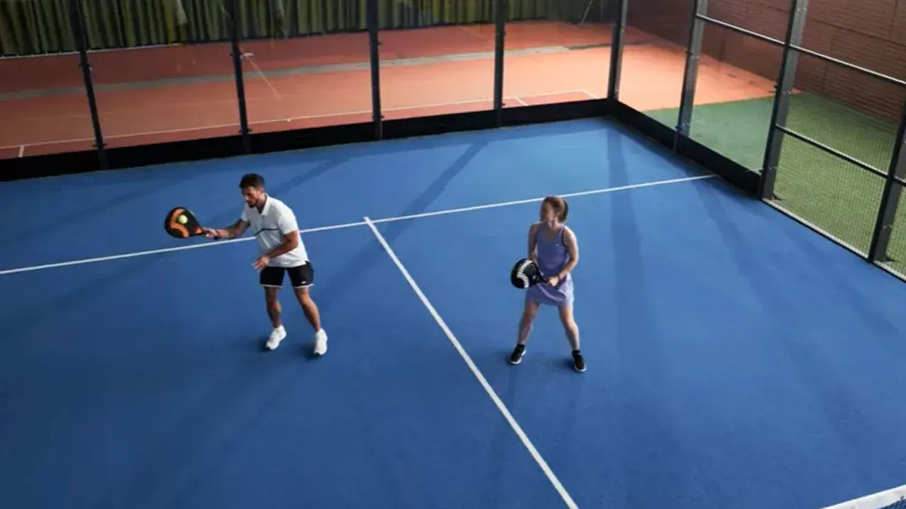 Coppia gioca a padel in campo blu. Lui colpisce la palla, lei attende il suo turno. Si può giocare a padel in 2.