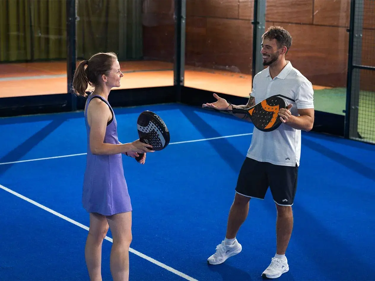 Coppia sorridente si prepara per una partita di padel misto, racchette in mano sul campo blu.