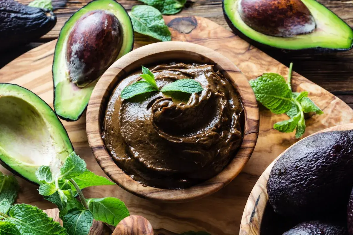 Una ciotola di legno contiene una deliziosa crema di avocado, guarnita con foglie di menta fresca. Avocadi maturi e foglie verdi circondano la ciotola.