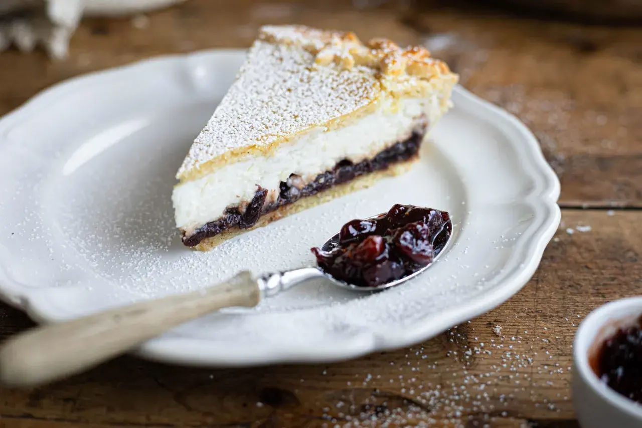 Una fetta di crostata ricotta e visciole, cosparsa di zucchero a velo, su un piatto bianco. Un cucchiaino pieno di visciole confettate accanto.