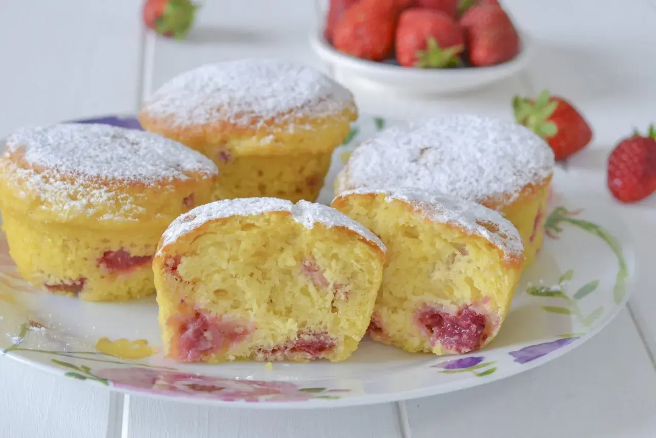 Morbidi muffin alle fragole, cosparsi di zucchero a velo, su un piatto fiorito. Uno è tagliato a metà, rivelando il ripieno succoso.