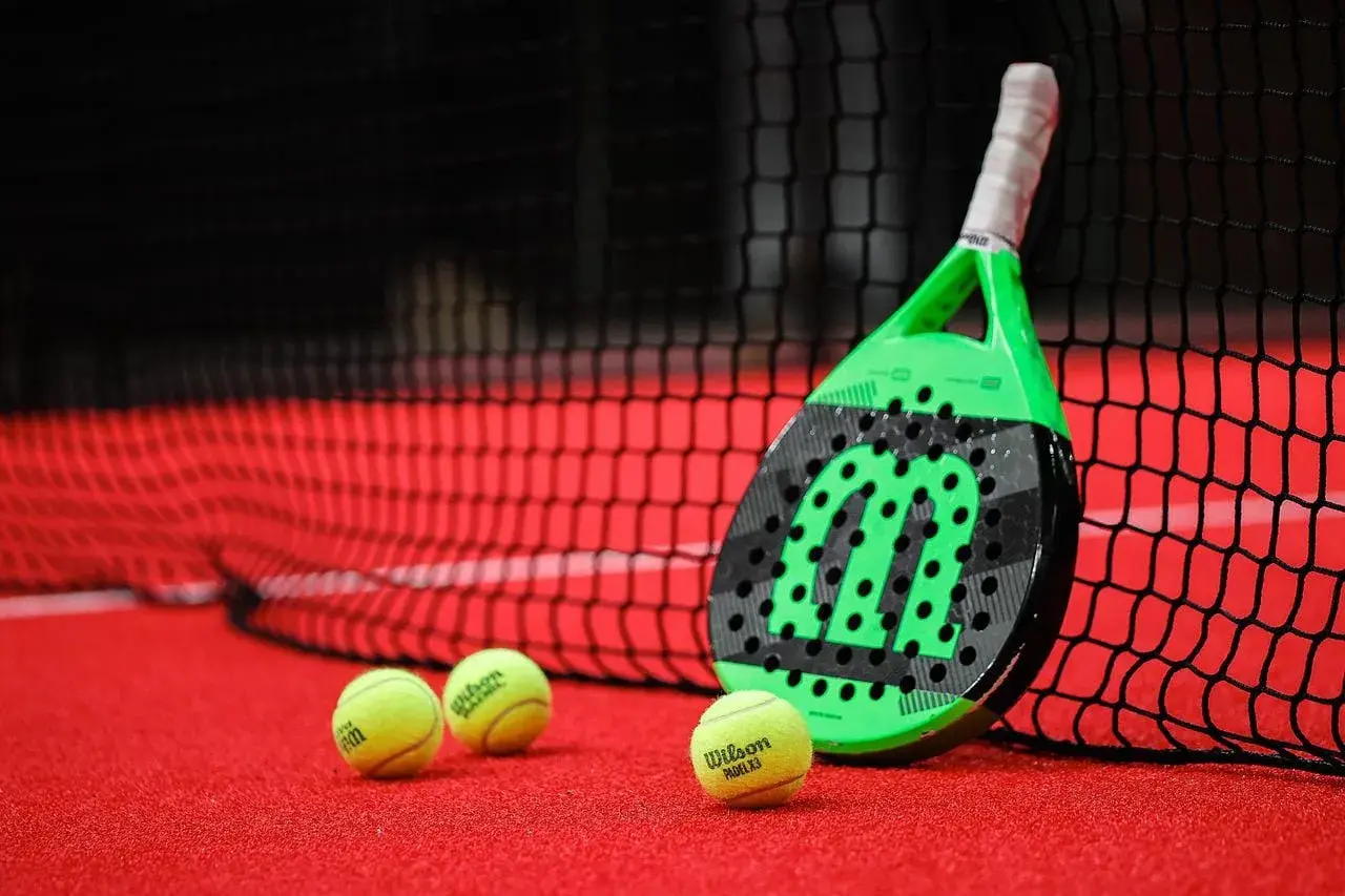 Racchetta da padel verde e nera appoggiata alla rete, con palline gialle sul campo rosso. Quanto dura una racchetta da padel? Dipende dall'uso!