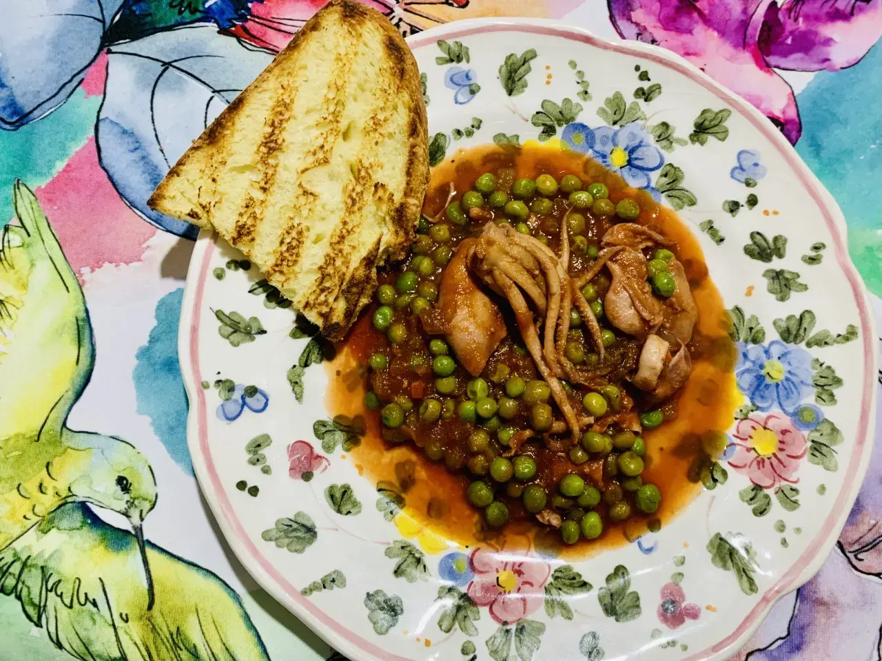 Piatto di totani con piselli in umido, accompagnato da una fetta di pane abbrustolito, su un piatto decorato con fiori e uccelli.
