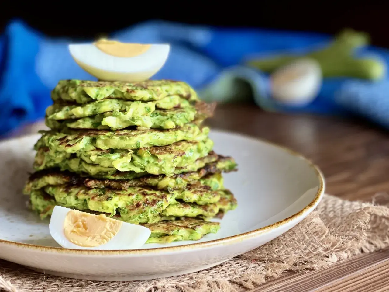Una pila di rosti di zucchine dorati, guarniti con spicchi di uovo sodo, su un piatto bianco.
