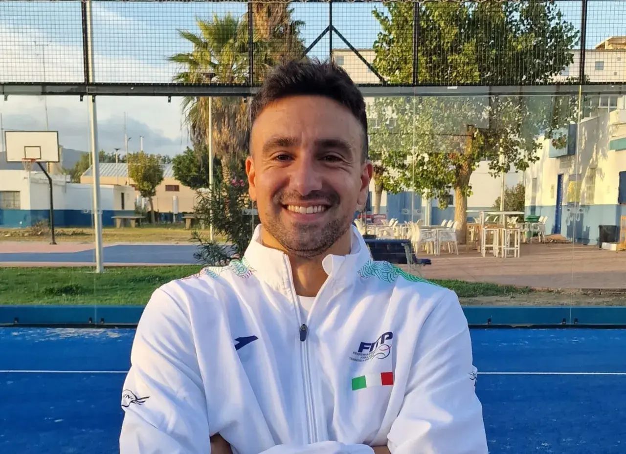Aris Patiniotis sorride con le braccia conserte su un campo da padel blu. Indossa una giacca bianca con logo e bandiera italiana.