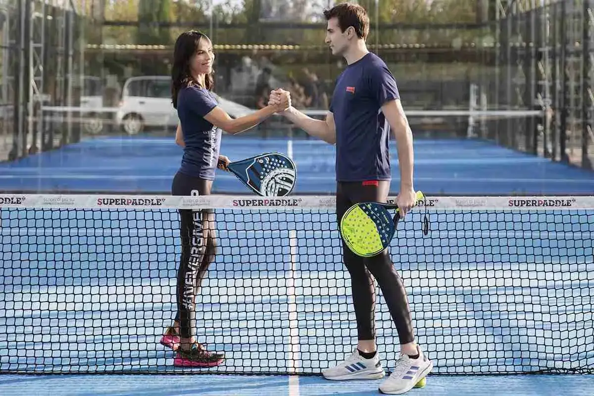 Coppia si stringe la mano su campo da padel, pronti a giocare. Lei indossa leggings con scritta, lui pantaloncini. Abbigliamento ideale per giocare a padel.