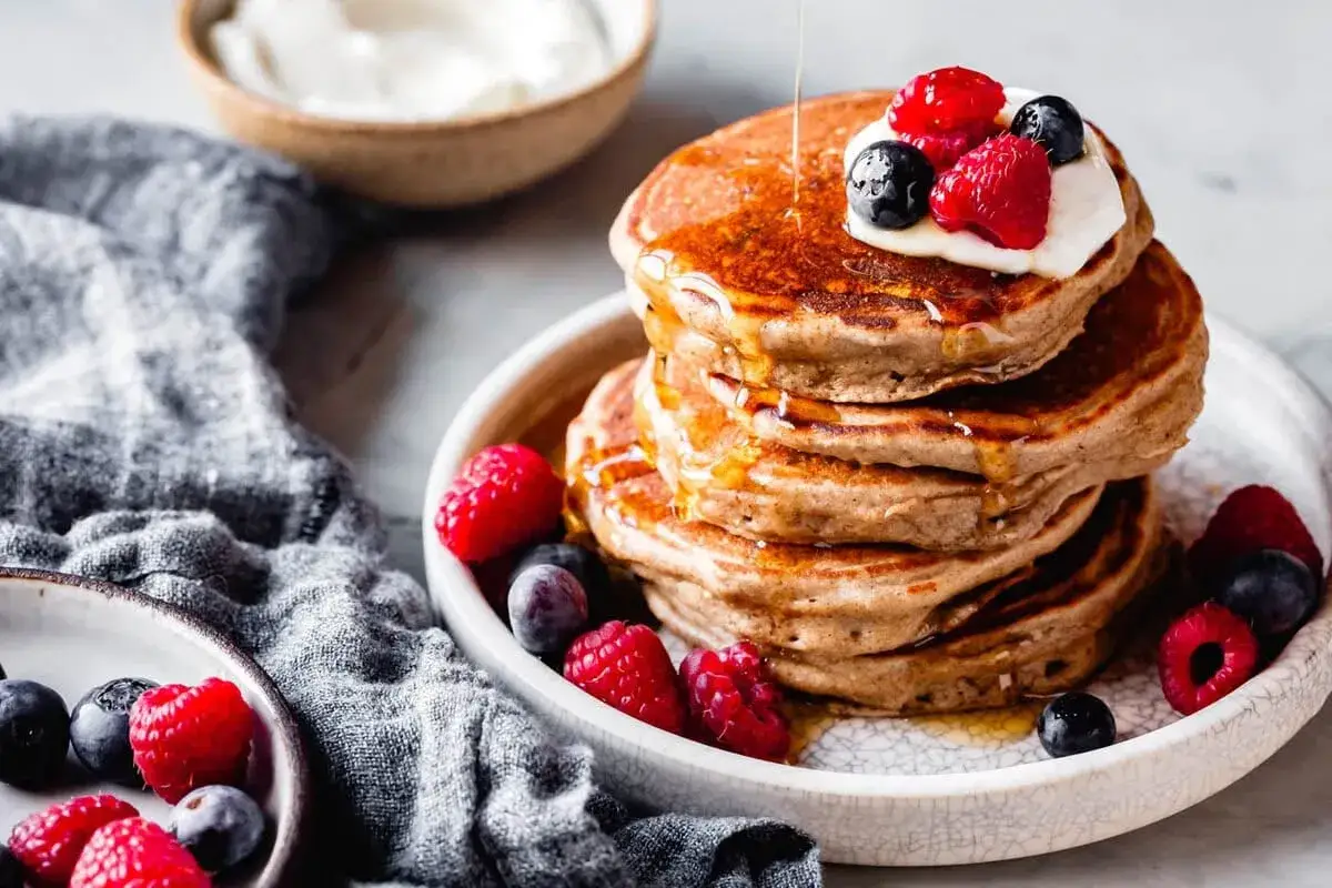 Pancakes con farina di avena, guarniti con panna, lamponi e mirtilli, inzuppati nello sciroppo d'acero.