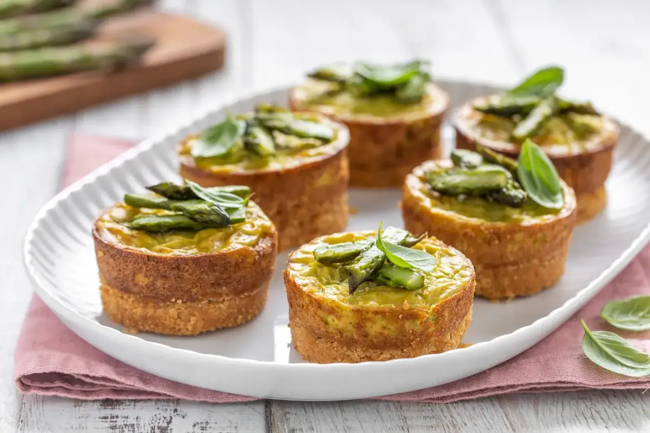 Sei tortini di asparagi, guarniti con germogli freschi e basilico, su un piatto bianco.