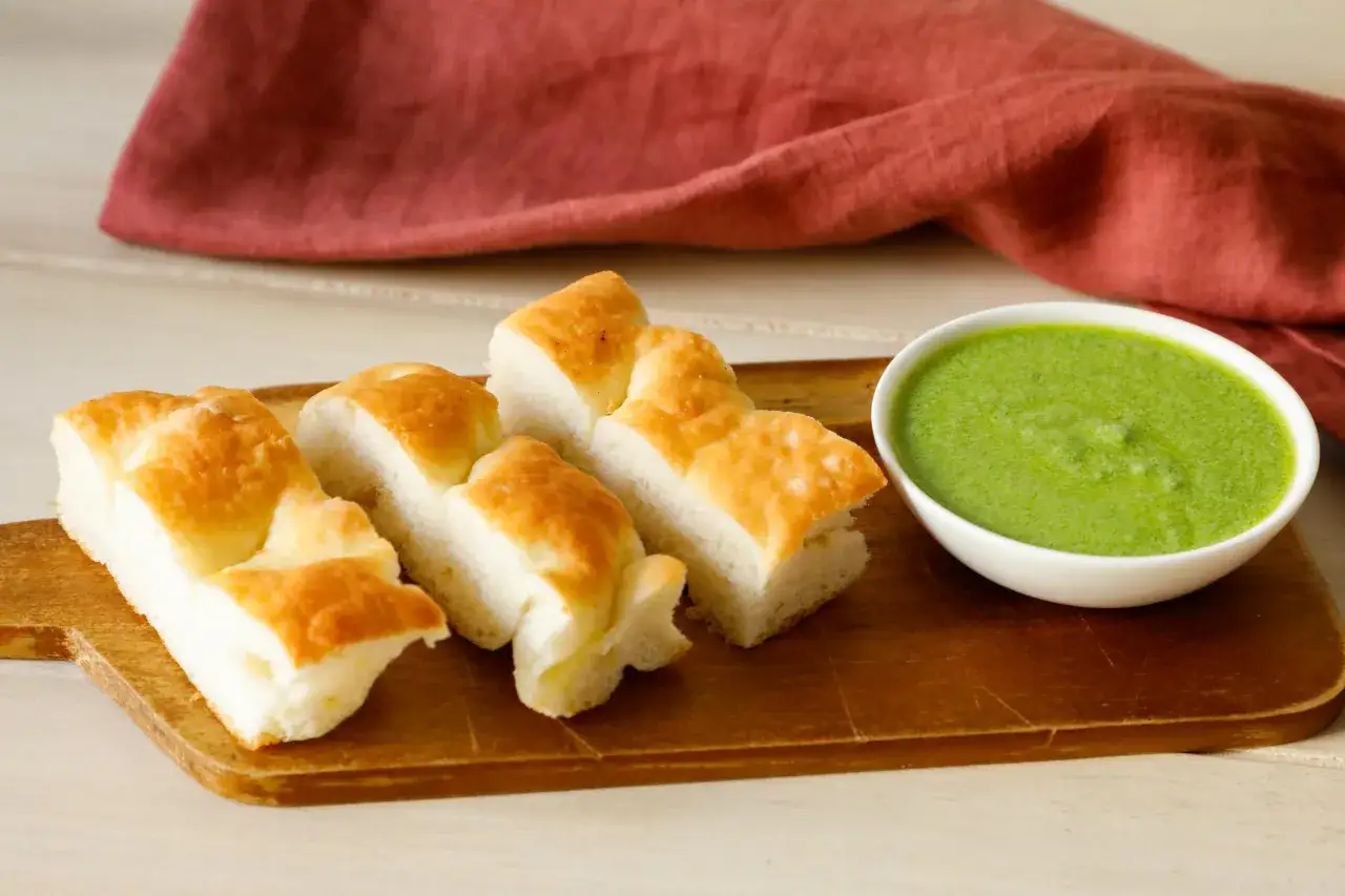 Focaccia tagliata a pezzi su tagliere di legno, accanto a una ciotola di pesto di salvia verde brillante.