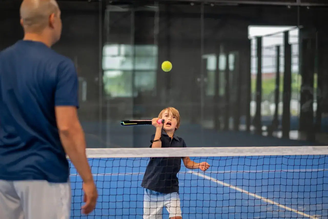 Un bambino, Nicola Bini, colpisce una palla da padel con entusiasmo, mentre un uomo lo osserva.