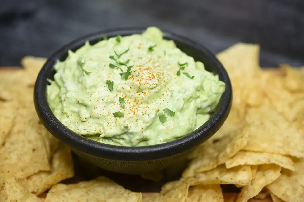 Una ciotola di cremosa crema di avocado, guarnita con prezzemolo e paprika, circondata da croccanti tortilla chips.