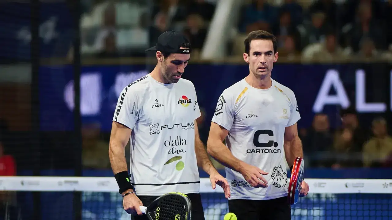 Due giocatori di padel, uno con un cappellino, si guardano pensierosi in campo. Il loro abbigliamento è sponsorizzato, con loghi come