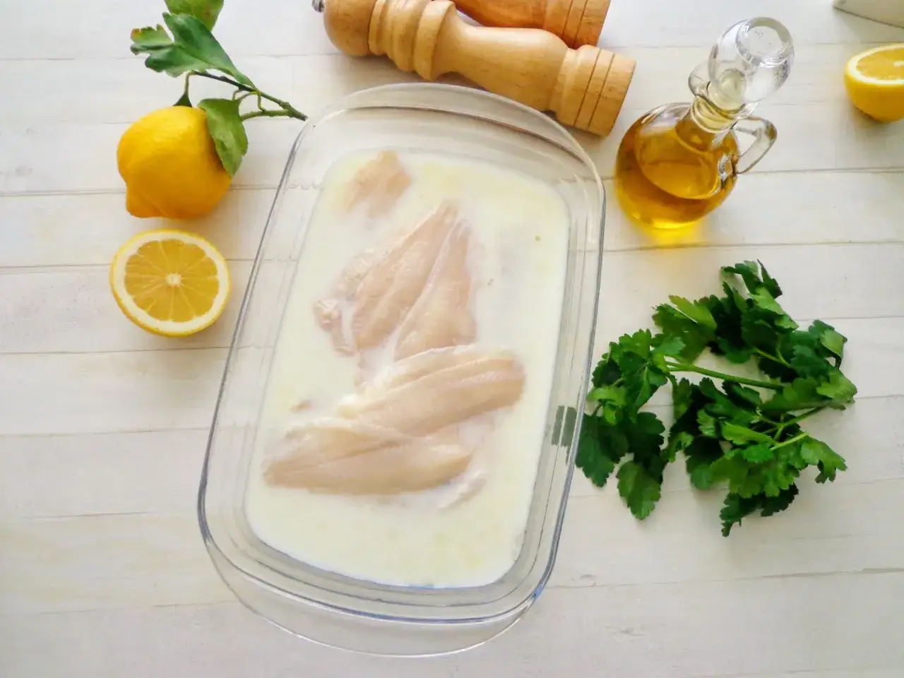 Filetti di platessa in latte, pronti per la mugnaia. Limone, olio e prezzemolo completano la scena.