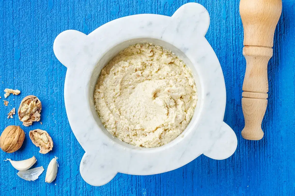 Mortai con pestello e ingredienti per preparare un cremoso pesto di noci su fondo blu.