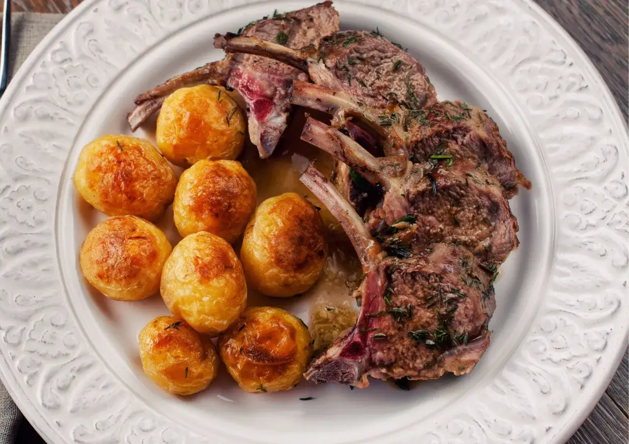 Costolette di agnello al forno, succulente e dorate, servite con patate novelle arrosto su un piatto bianco decorato.