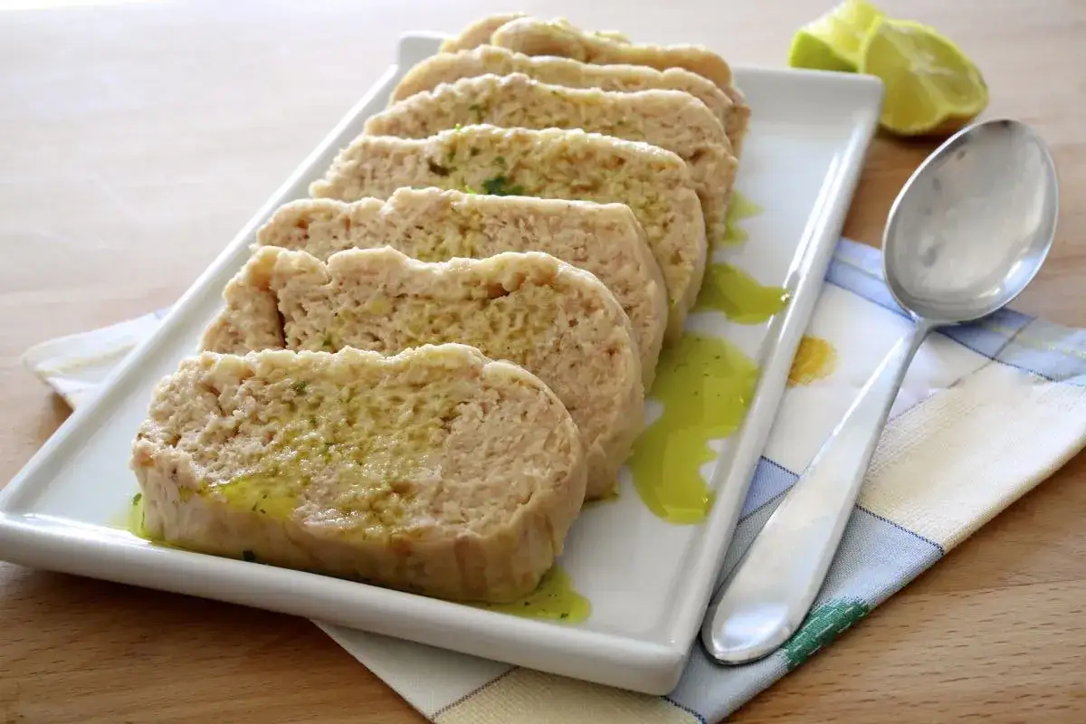 Fette di polpettone di tonno su un piatto bianco, condite con olio e erbe. Accanto, un cucchiaio e spicchi di lime.