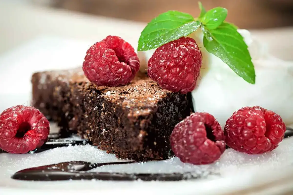 Una fetta di torta all'acqua al cacao, spolverata di zucchero a velo e cacao, guarnita con lamponi freschi e una fogliolina di menta.