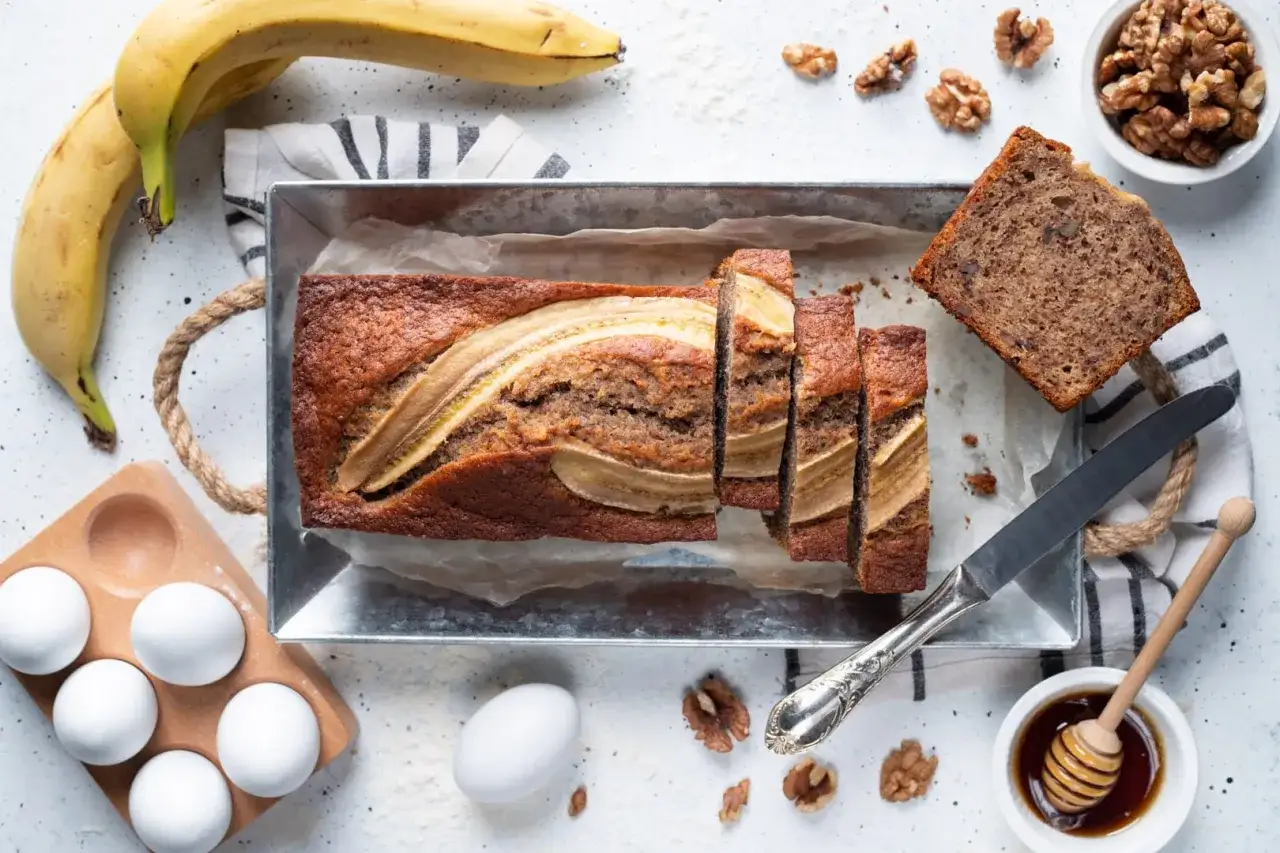 Fetta di banana bread con noci, accanto a banane mature, uova e miele.
