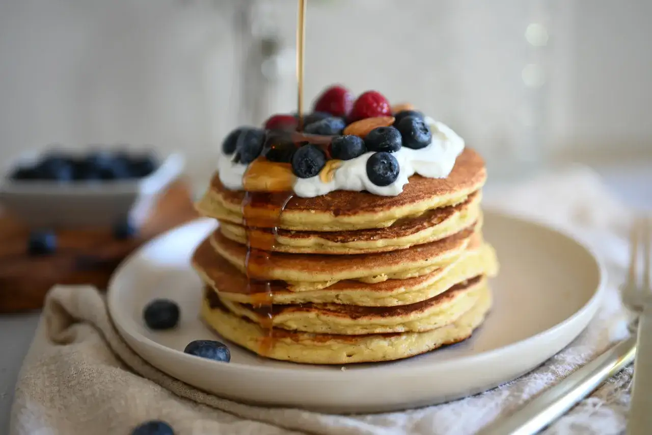 Pancake senza uova con yogurt, guarniti con mirtilli, lamponi e mandorle, con sciroppo d'acero che cola.
