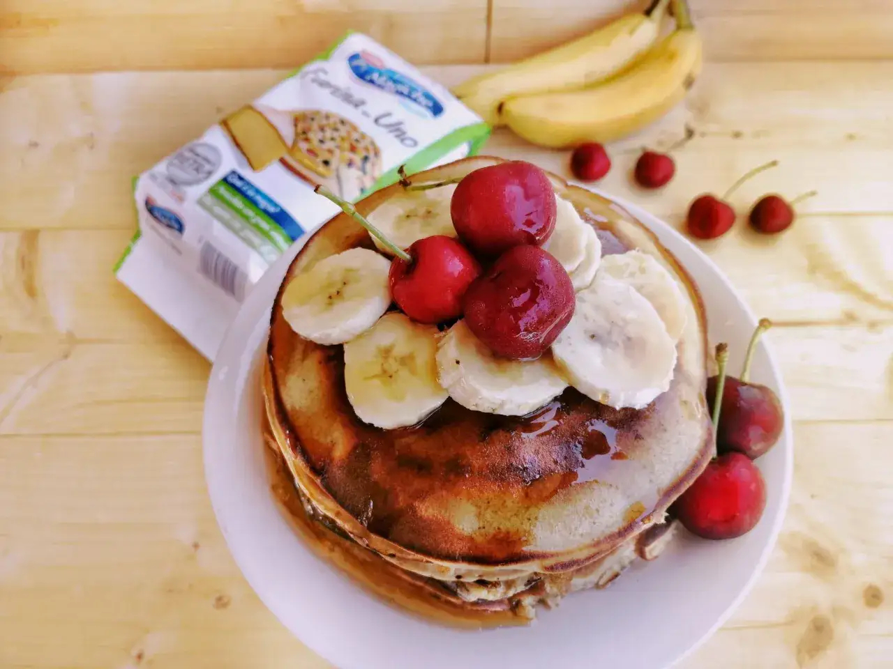 Pancake alla banana con ciliegie e sciroppo, pronti per una colazione golosa. Sullo sfondo, banane e una confezione di farina.