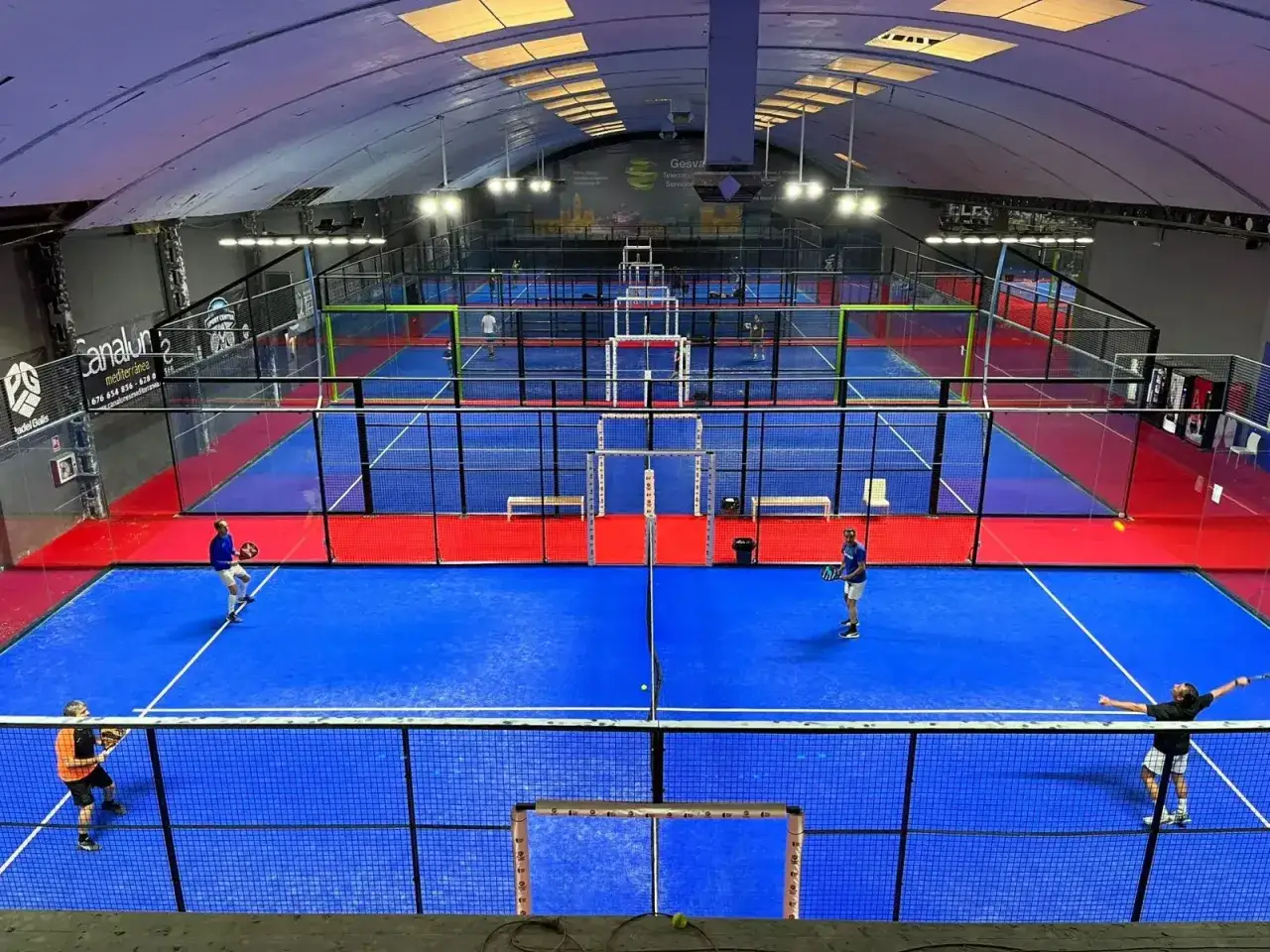 Campi da padel in Spagna, con giocatori che si sfidano su campi blu e rossi sotto un soffitto curvo illuminato.