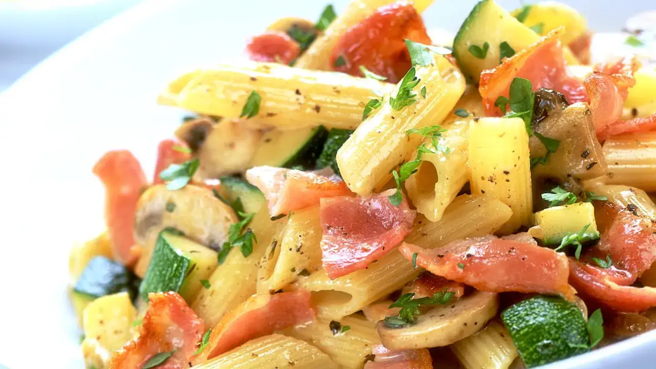 Penne con zucchine, pancetta croccante e funghi, guarnite con prezzemolo fresco. Un piatto saporito e colorato.