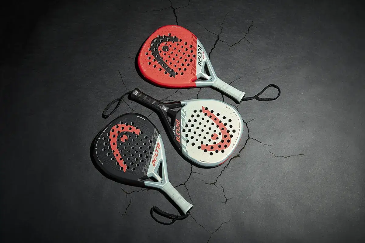 Tre racchette da padel Delta, una rossa, una bianca e una nera, giacciono su una superficie scura e screpolata.